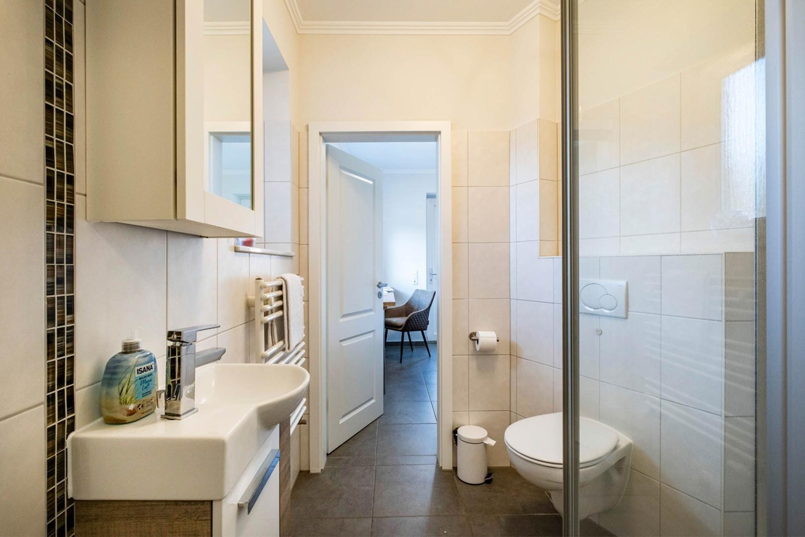 Badezimmer mit Waschbecken, Toilette und Dusche. Blick in angrenzenden Raum.
