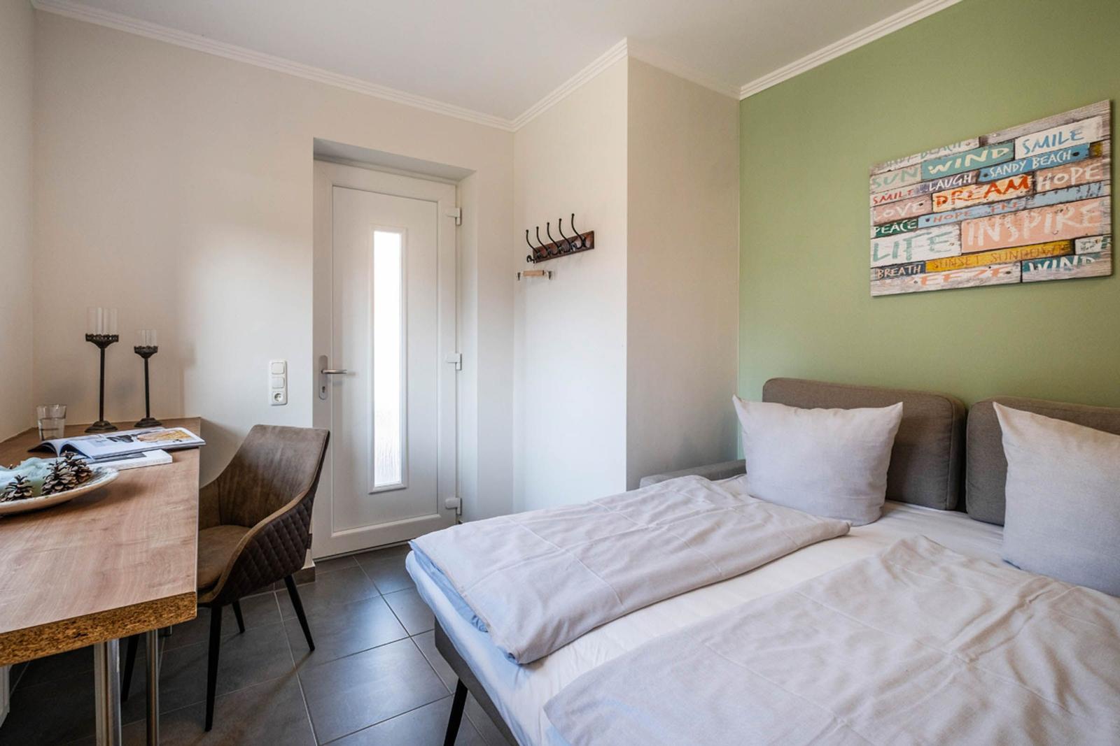 Schlafzimmer mit Bett, Tisch, Stuhl und Wandbild. Tür und Hakenwand sind sichtbar.