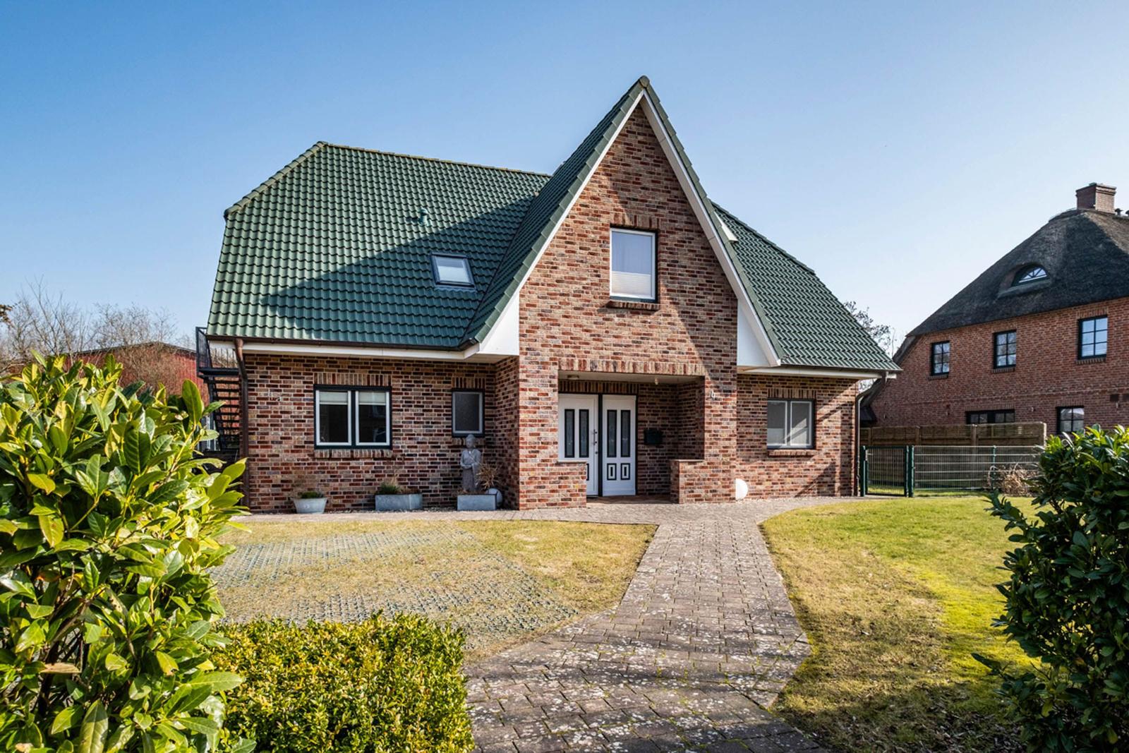 Ein zweistöckiges Haus mit rotem Backstein und grünem Dach. Vorgarten mit Pflanzen und Steintreppe.