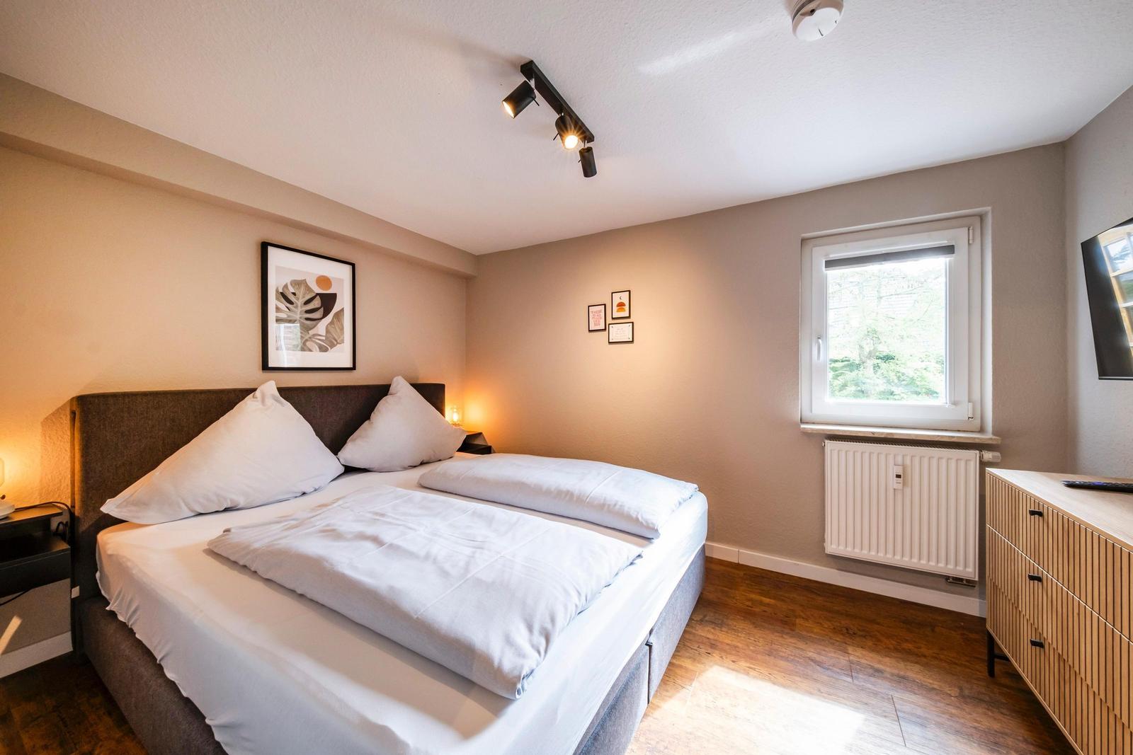 Schlafzimmer mit Bett, Fenster und Schrank. Wandbild und Beleuchtung sichtbar.