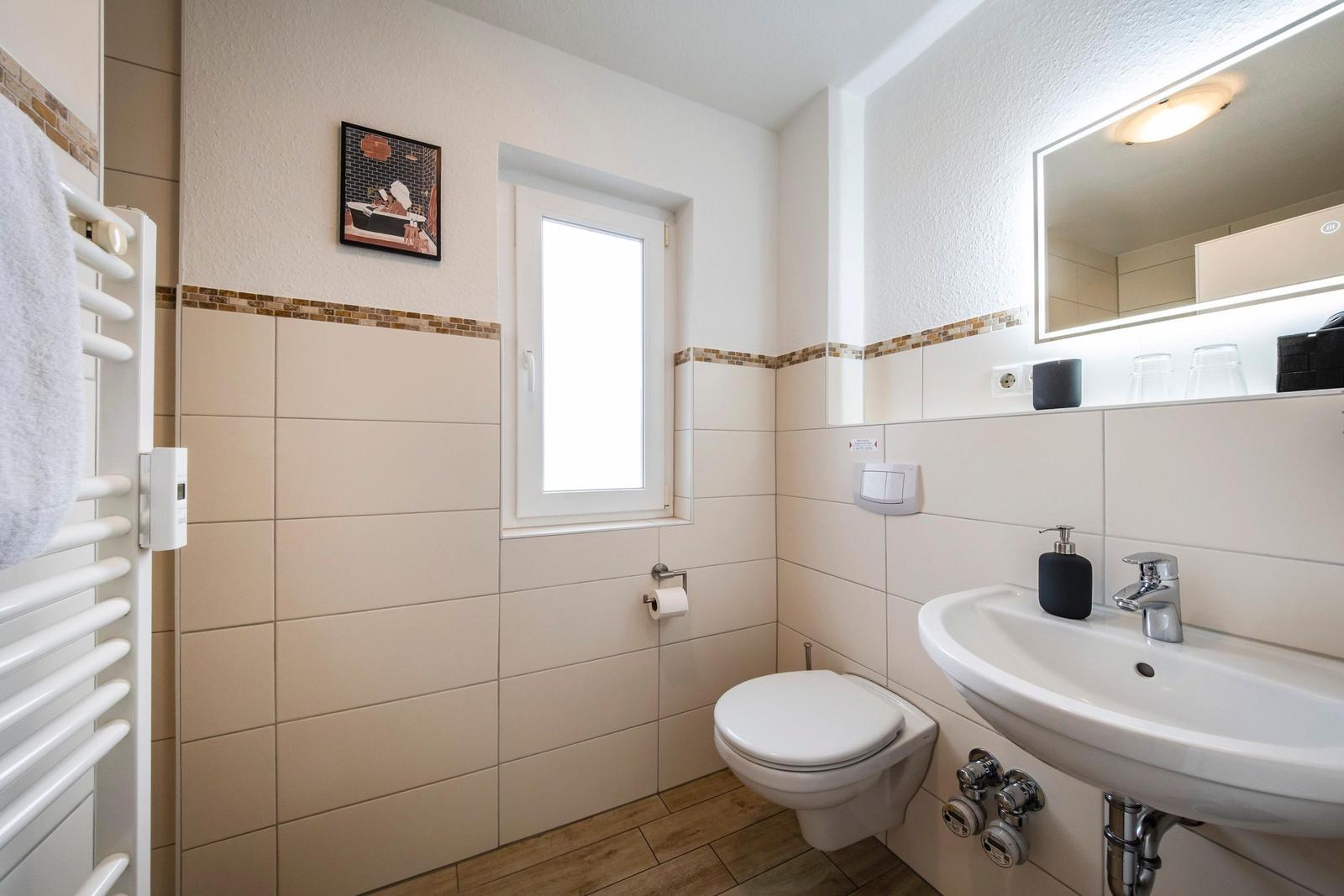 Badezimmer mit Toilette, Waschbecken und Fenster. Wandfliesen und Spiegel.