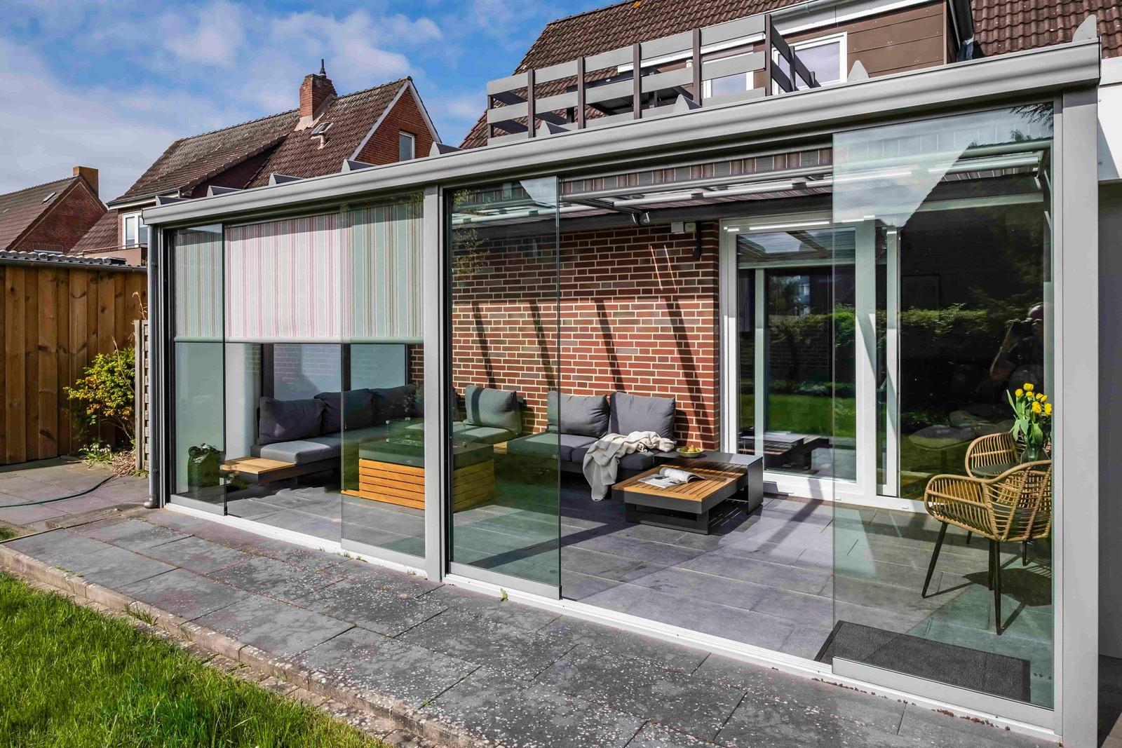 Glasveranda mit Sitzgruppe und Tisch. Hintergrund: Backsteinmauer und Garten.