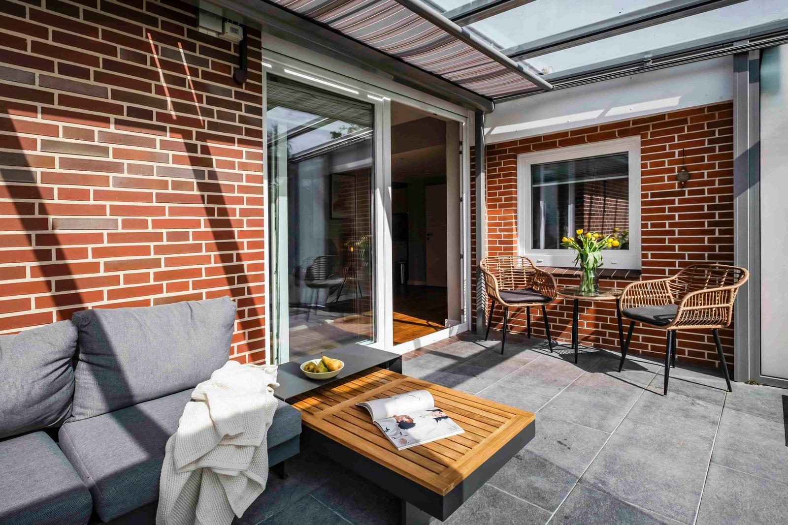 Terrasse mit Sofa, Tisch und Stühlen. Roter Backstein, Glaswand und Sonnenschutz.