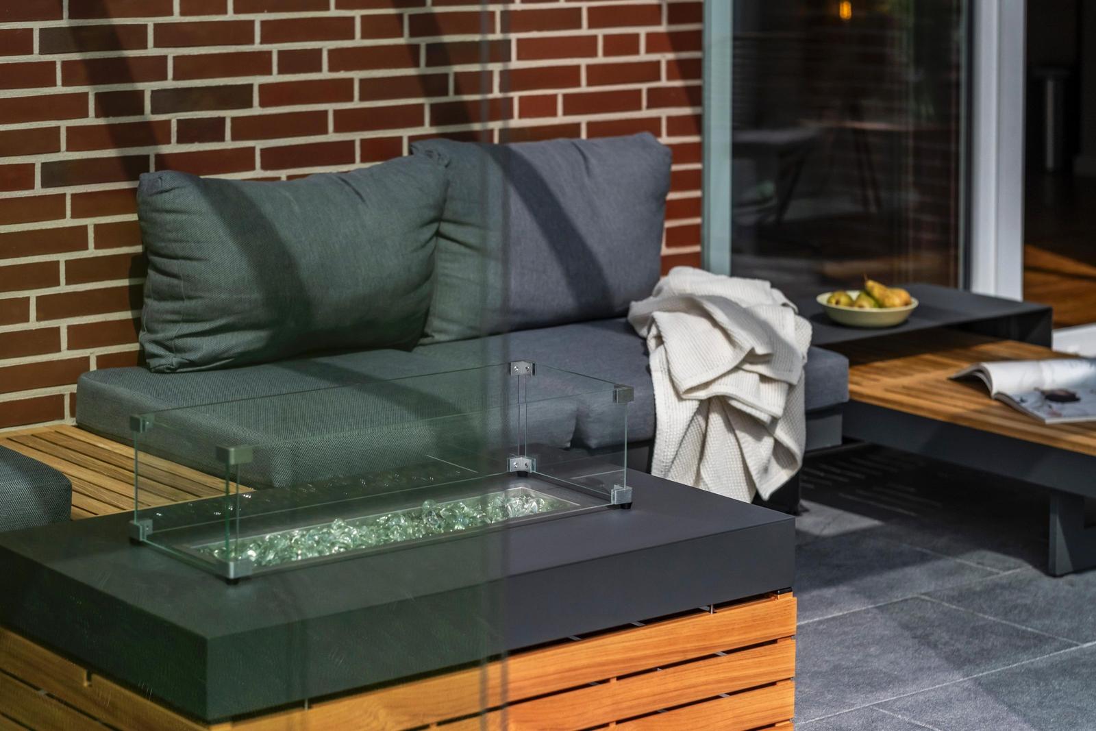 Außenbereich mit Sofa, Glasfeuer und Holztisch. Hintergrund: Backsteinmauer und Glastür.