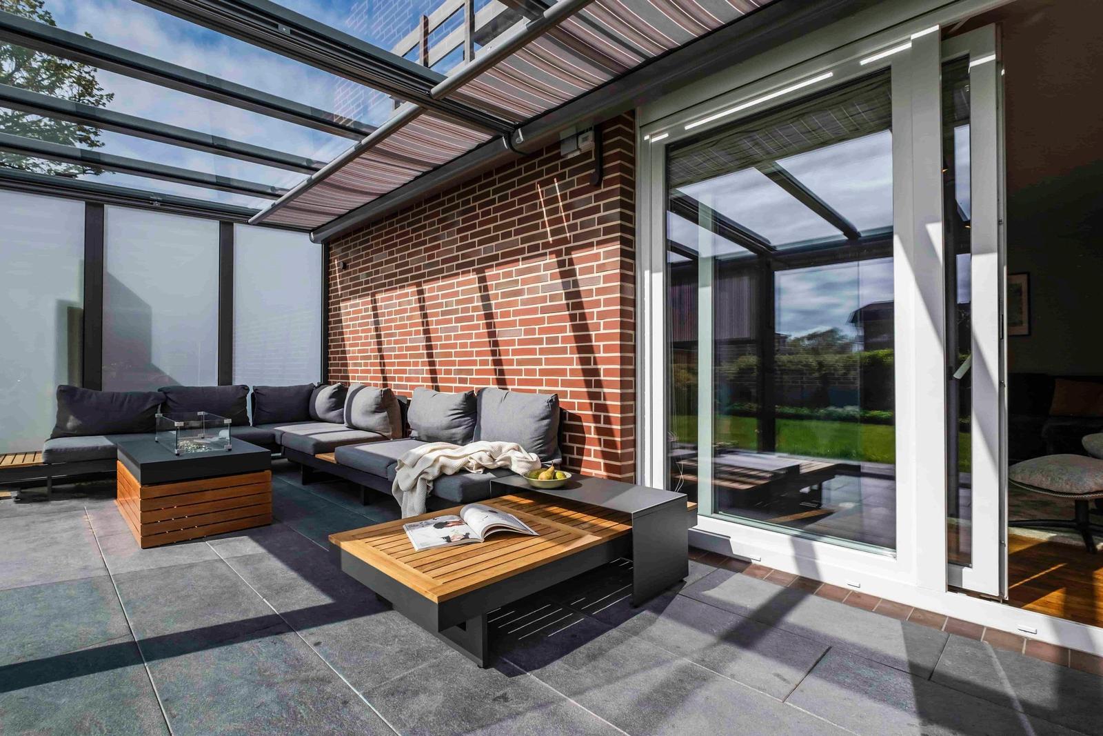 Terrasse mit Sitzgruppe, Holztisch und Glasdach. Roter Backsteinwand und Schiebetür.