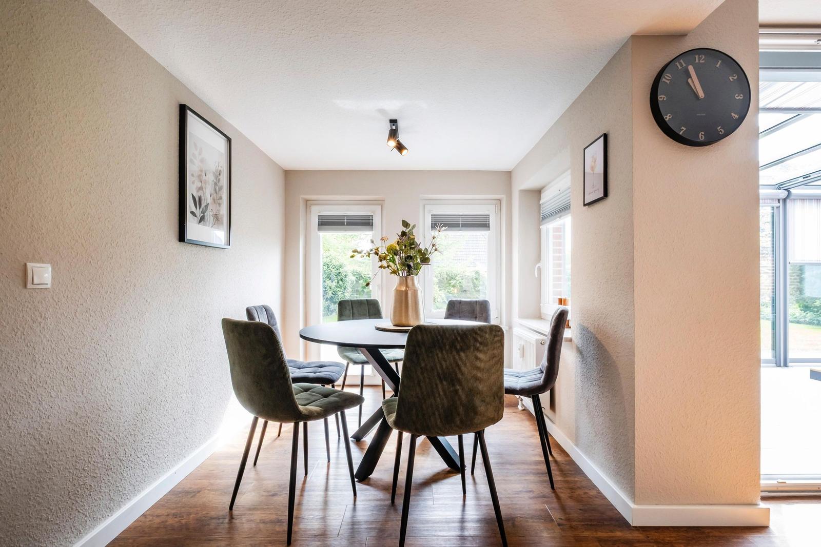 Esszimmer mit runder Tisch und Stühlen, Wanduhr und Fenstern.