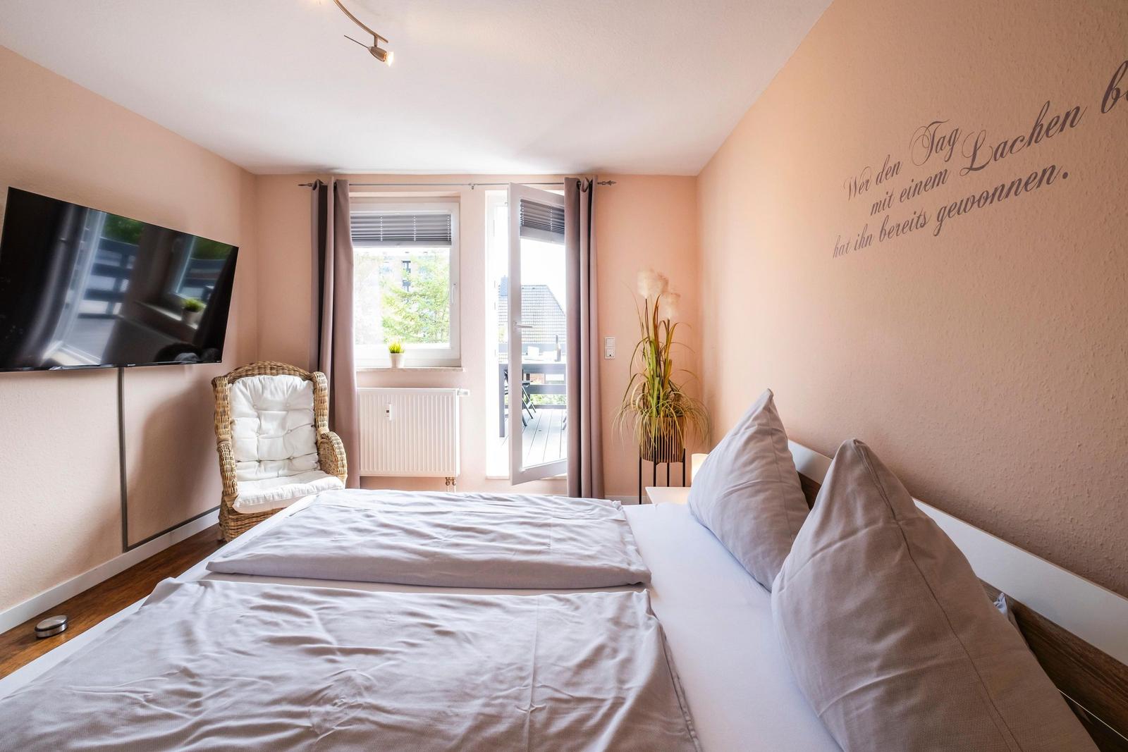 Schlafzimmer mit Bett, Fernseher und Balkontür. Wanddekoration und Pflanze sichtbar.
