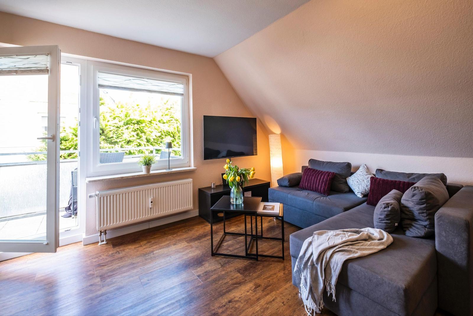 Wohnzimmer mit Sofa, Fernseher und Balkontür. Holzboden und schräge Wand.