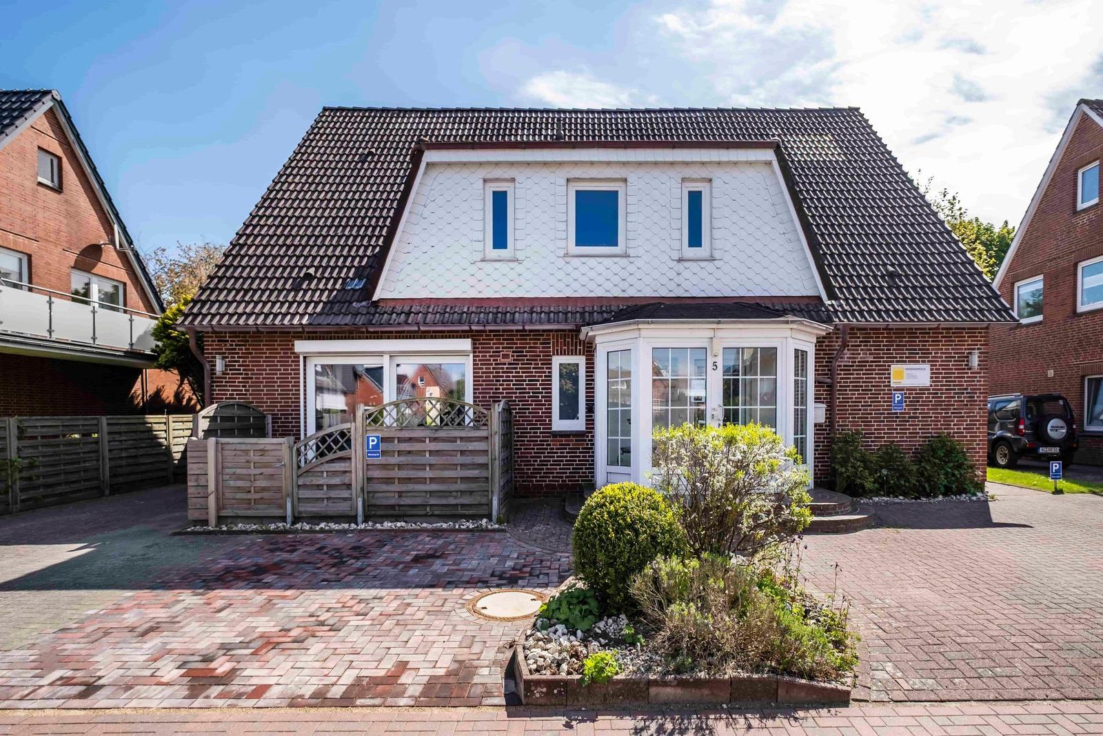 Haus mit braunem Dach und rotem Backstein, weißer Veranda und gepflasterter Vorgarten.