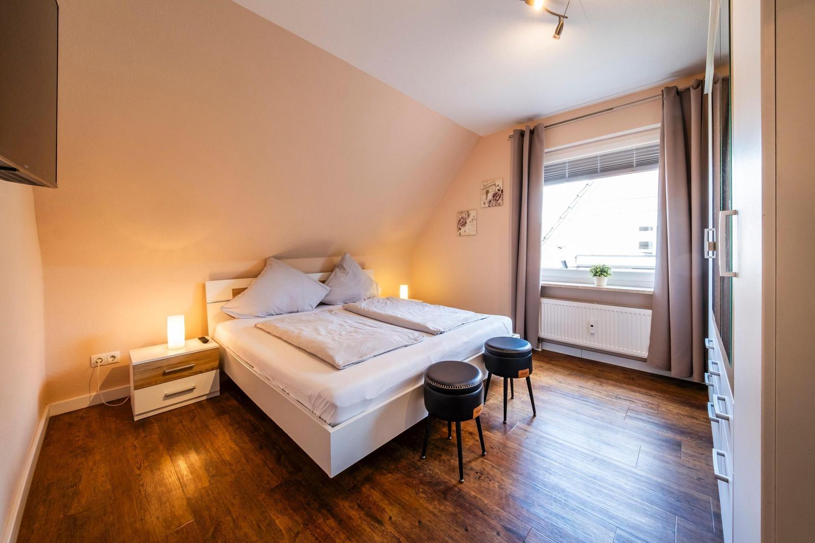 Schlafzimmer mit Doppelbett, Nachttisch, zwei Hocker und Fenster mit Vorhängen.