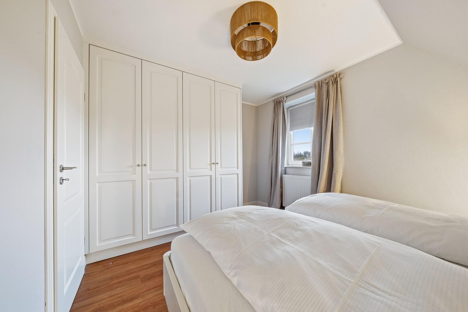 Schlafzimmer mit Doppelbett, weißen Schränken und Holzboden. Fenster mit Vorhängen und Deckenleuchte.