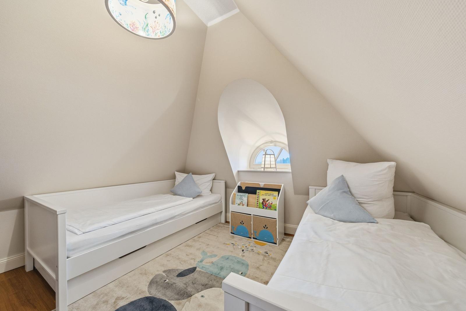 Zwei Einzelbetten in einem Dachzimmer mit Rundfenster und Kinderbuchregal.
