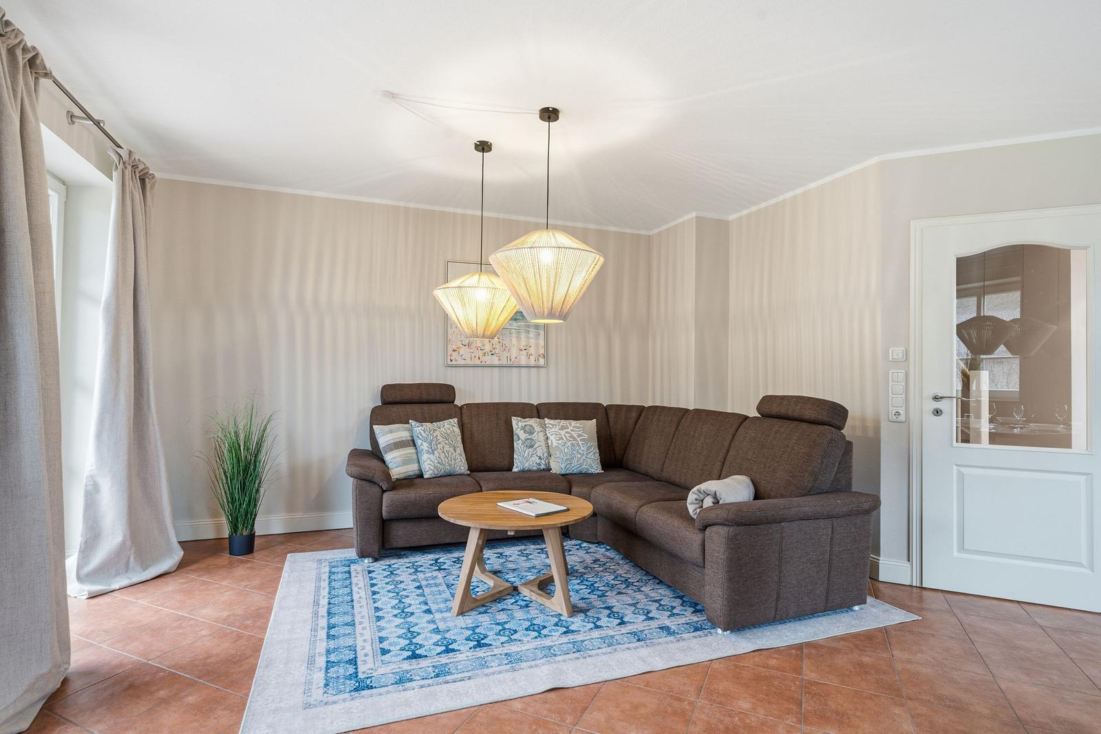 Wohnzimmer mit Ecksofa, runden Tisch und blauem Teppich auf Terrakottboden.