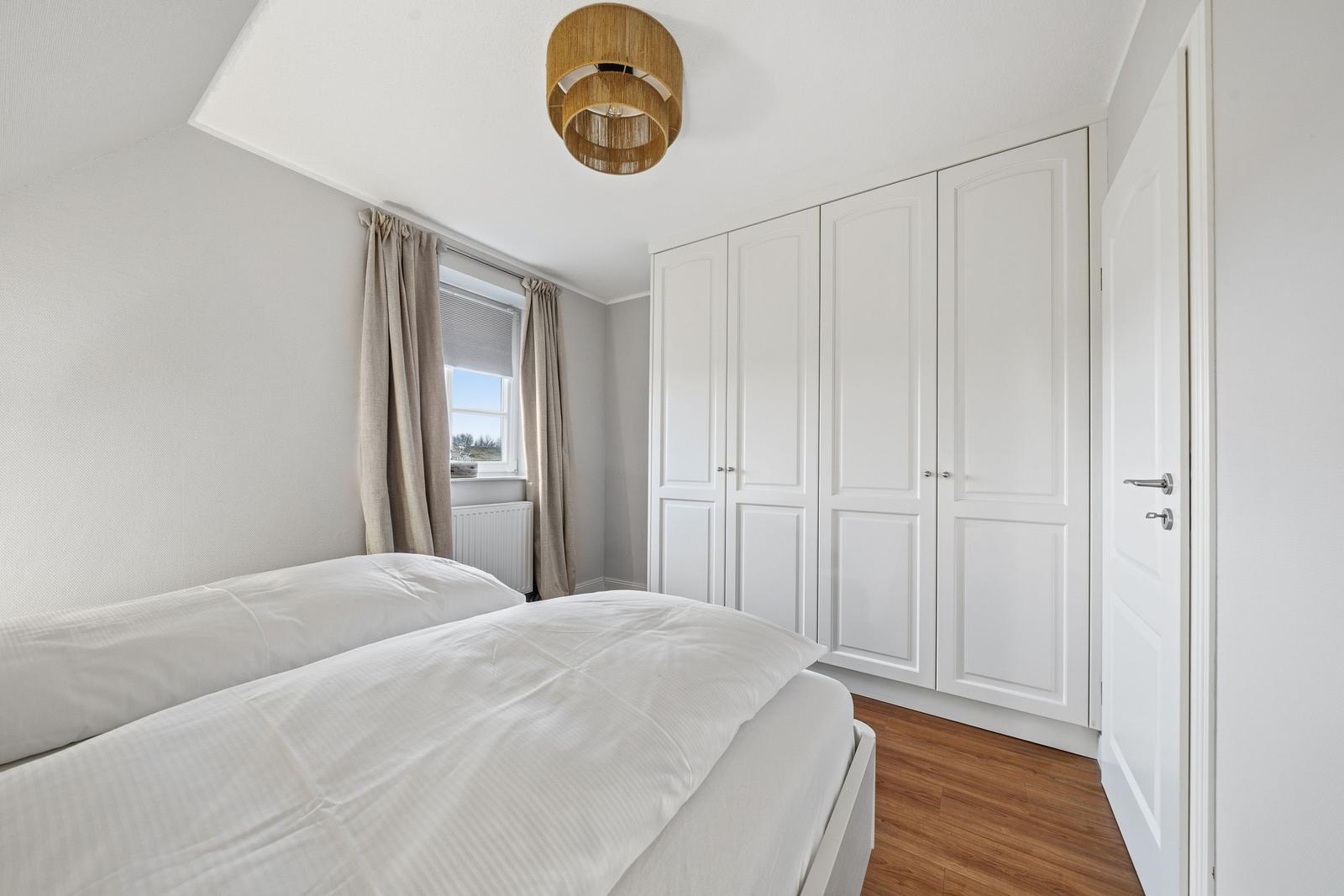 Schlafzimmer mit weißem Bett, Kleiderschrank und Fenster mit Vorhängen.