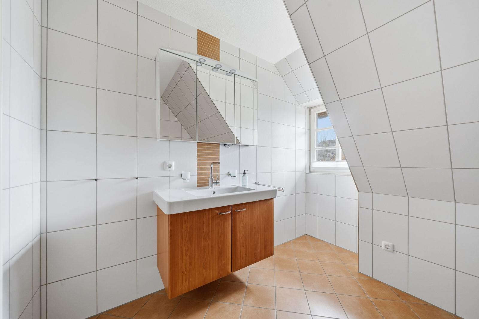 Badezimmer mit Waschbecken, Spiegel und Fenster. Wände und Boden sind gefliest.