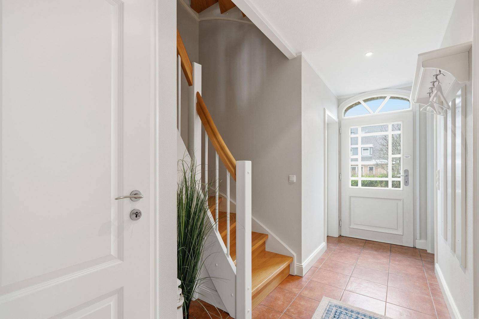 Hausflur mit Holztreppe, weißen Türen und Terrakottaboden. Fenster mit Bogen oben.