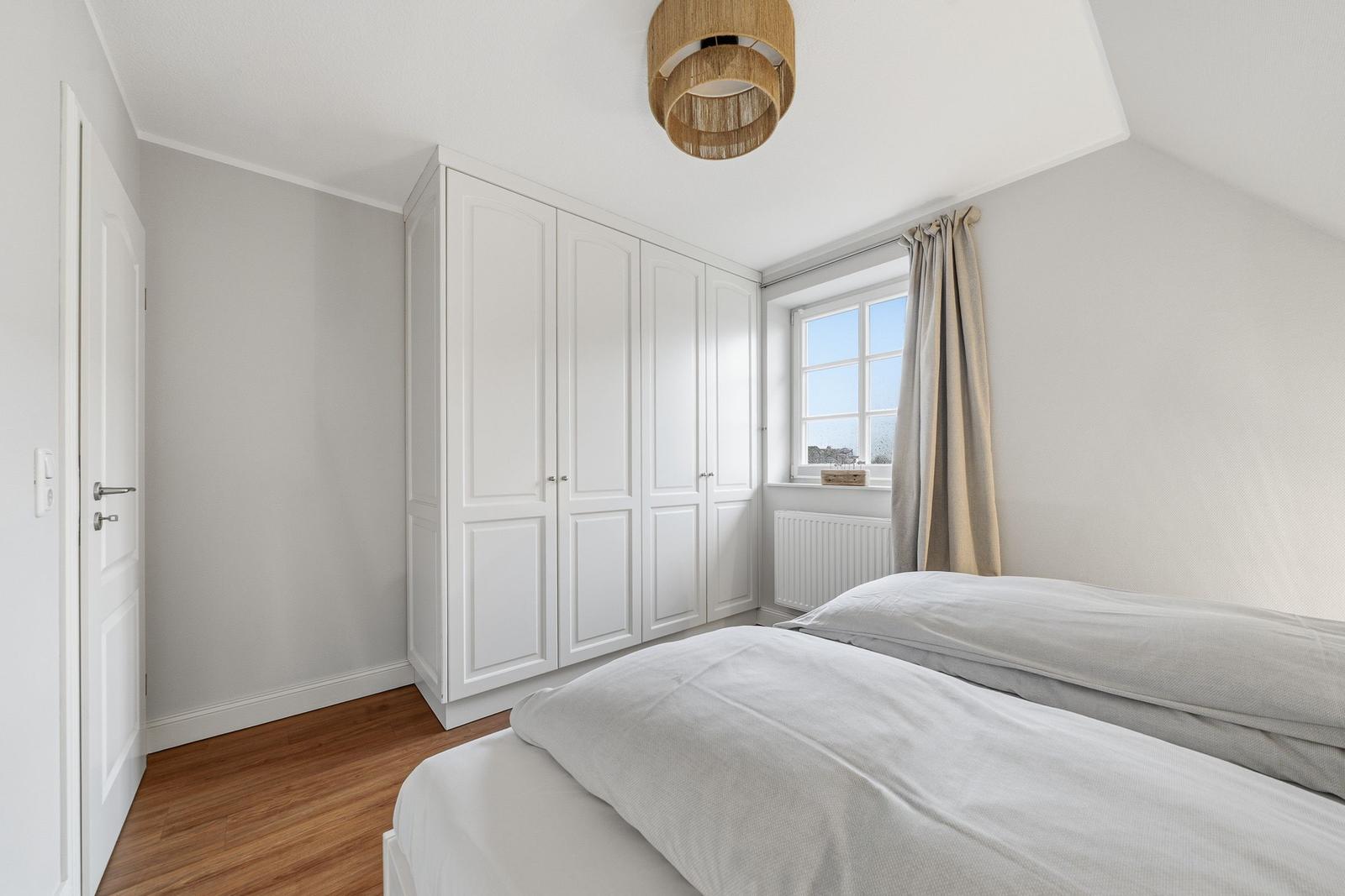 Schlafzimmer mit Doppelbett, weißem Schrank und Fenster mit Vorhängen.