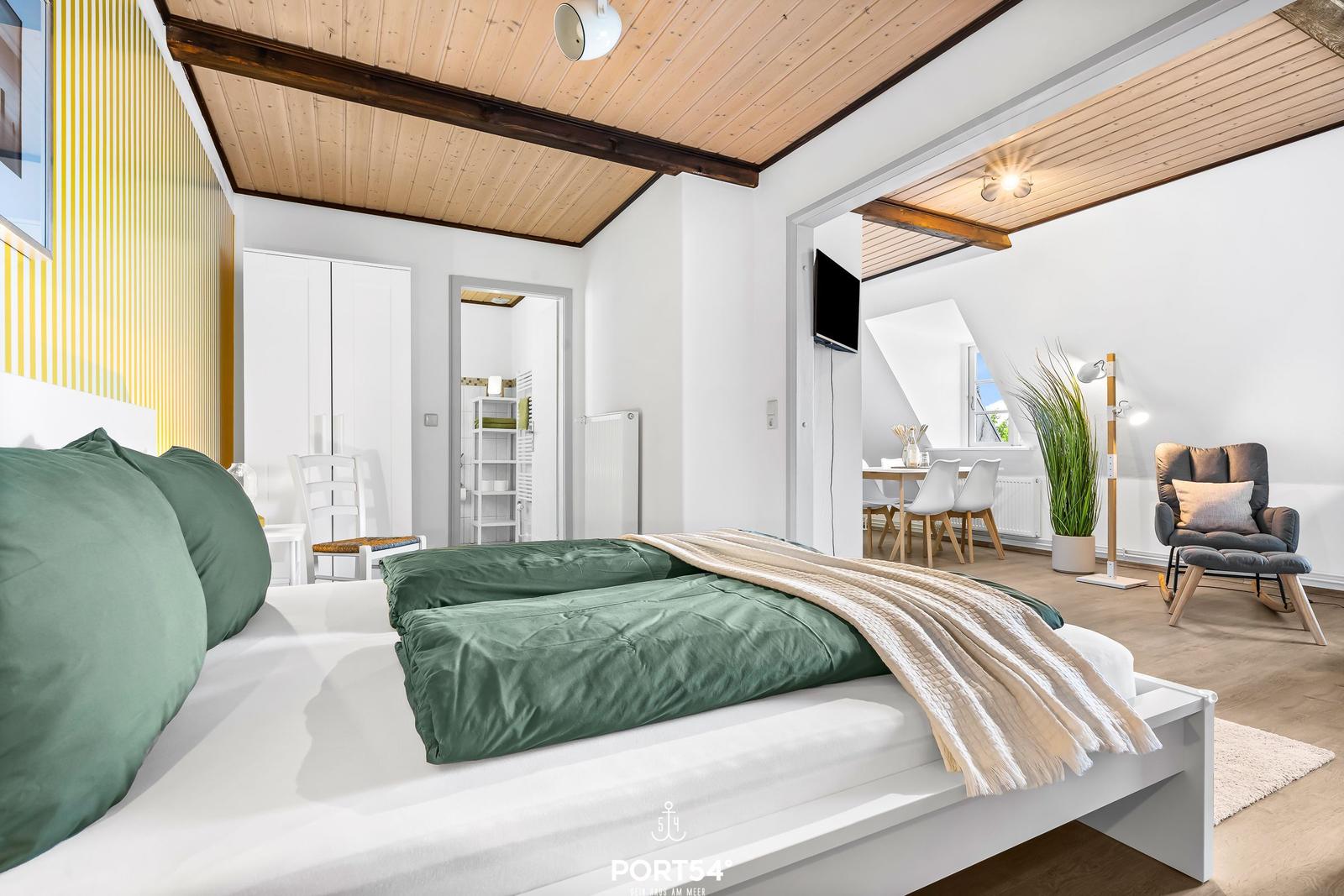 Schlafzimmer mit Bett, grüner Bettwäsche und Blick in Wohnbereich mit Sessel und Esstisch.