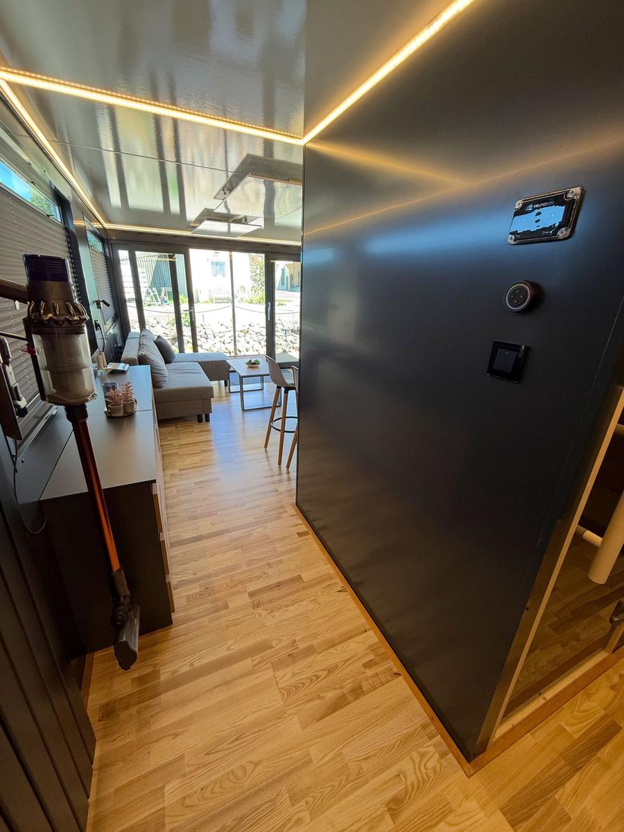Entrance area with wooden floor and modern lighting. In the background is the living room with sofa and table.