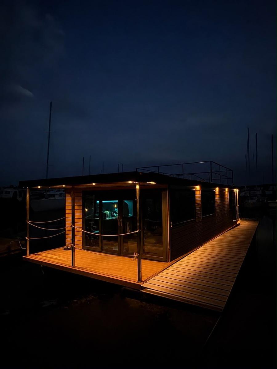 Modern houseboat with wooden deck and lit windows at night.