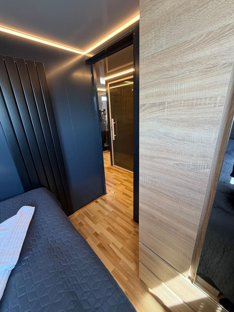 Bedroom with bed, wooden floor, and modern lighting. Door leads to adjacent room.