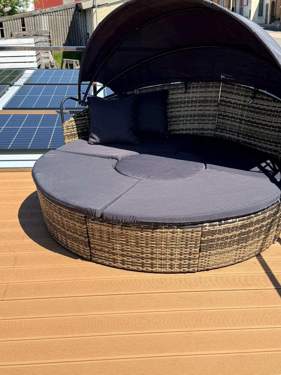 Round rattan lounge area with dark cushions and sunshade on terrace.