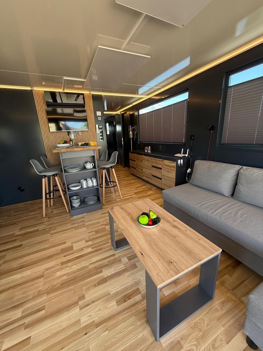 Living area with sofa, dining table, and kitchenette. Wooden floor and windows with blinds.