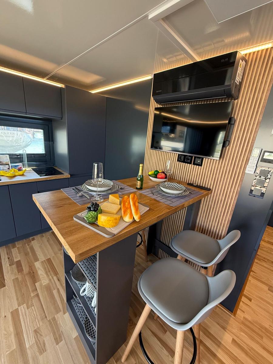 Kitchen area with wooden counter, stools, and provisions. Background: TV and sink.