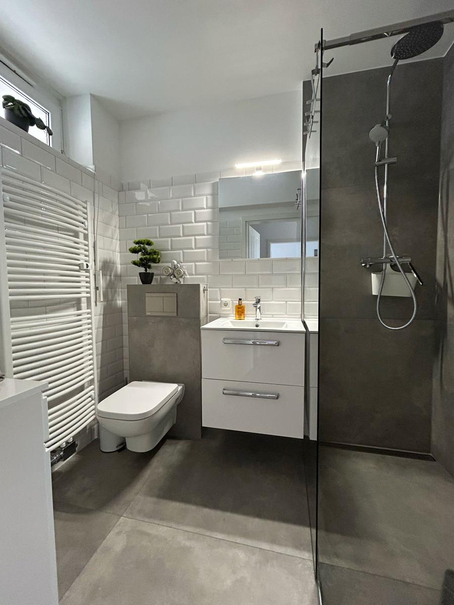 Badezimmer mit Dusche, Waschbecken und Toilette. Weiße Fliesen und graue Wandfliesen.