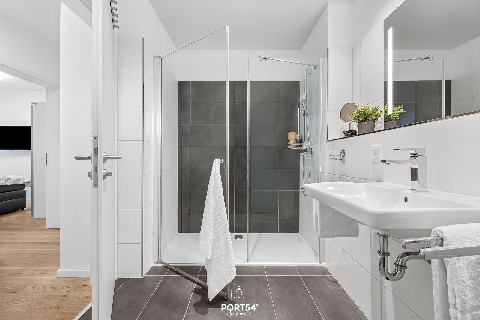Badezimmer mit Dusche, Waschbecken und Spiegel. Fliesen in Grau und Weiß.