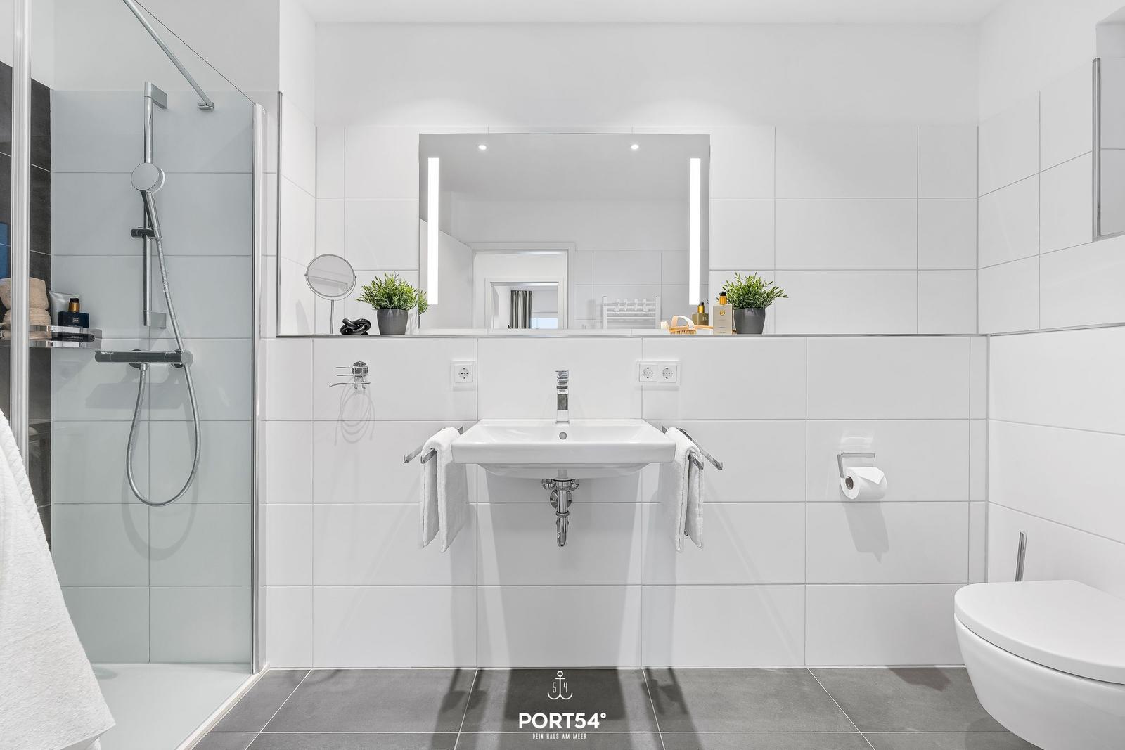 Badezimmer mit Dusche, Waschbecken und Toilette. Weiße Fliesen und Spiegel mit Beleuchtung.