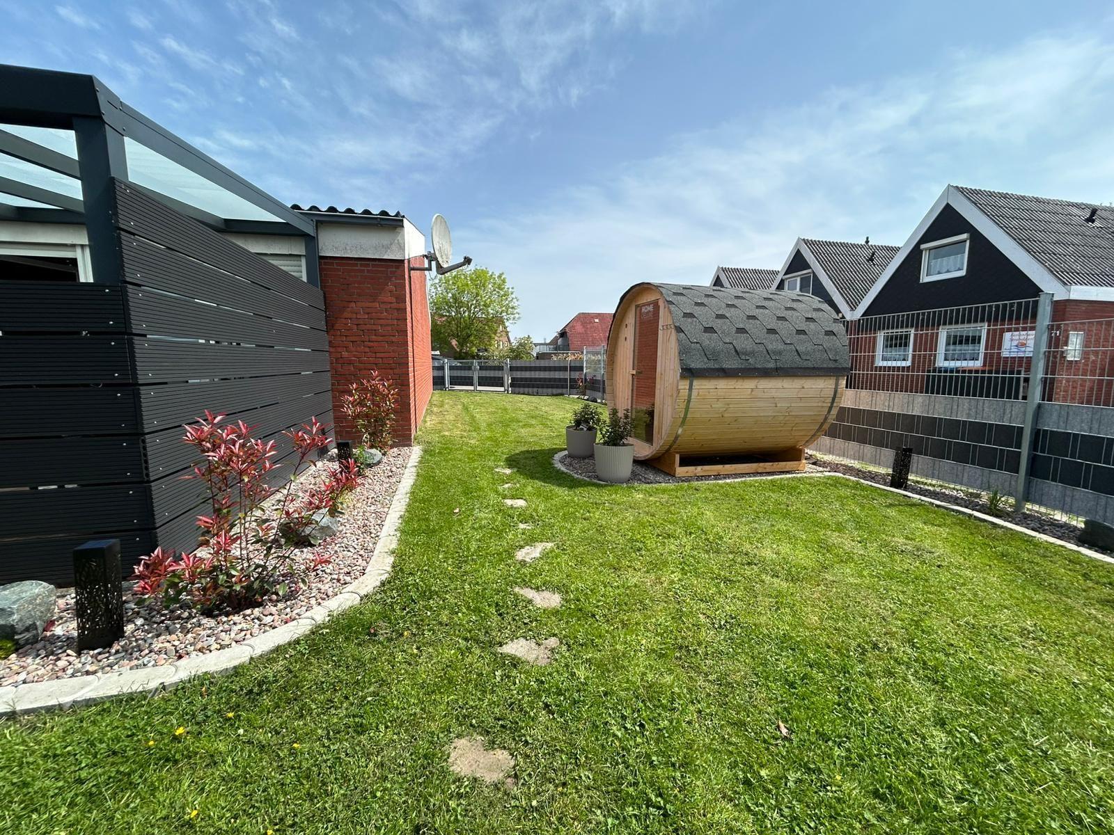 Backyard with sauna, lawn, and plants. Modern facade and fence visible.