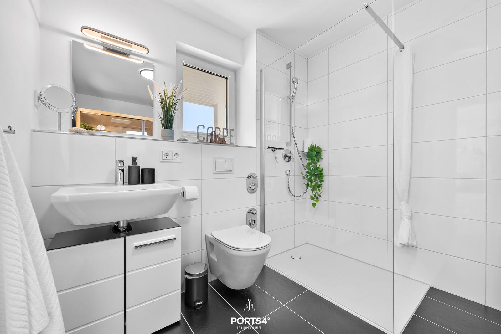 Badezimmer mit Dusche, Waschbecken und Toilette. Weiße Fliesen, schwarze Bodenfliesen.