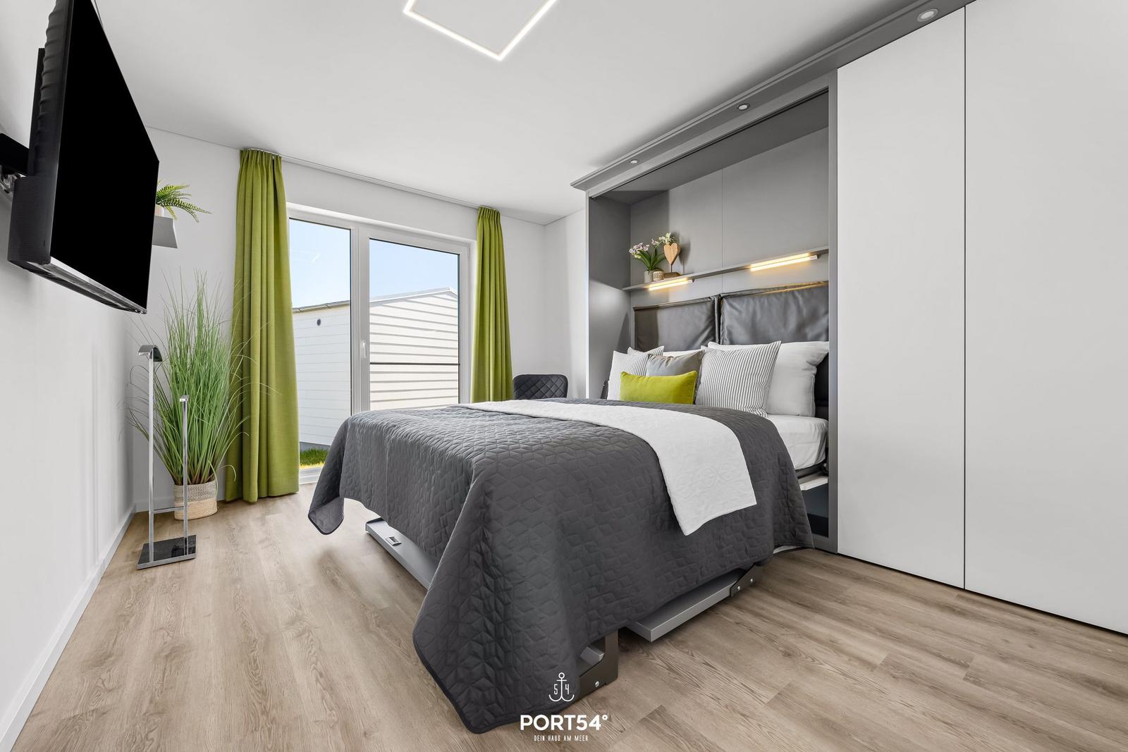 Schlafzimmer mit Bett, Schrank und TV. Grüne Vorhänge und Holzboden.