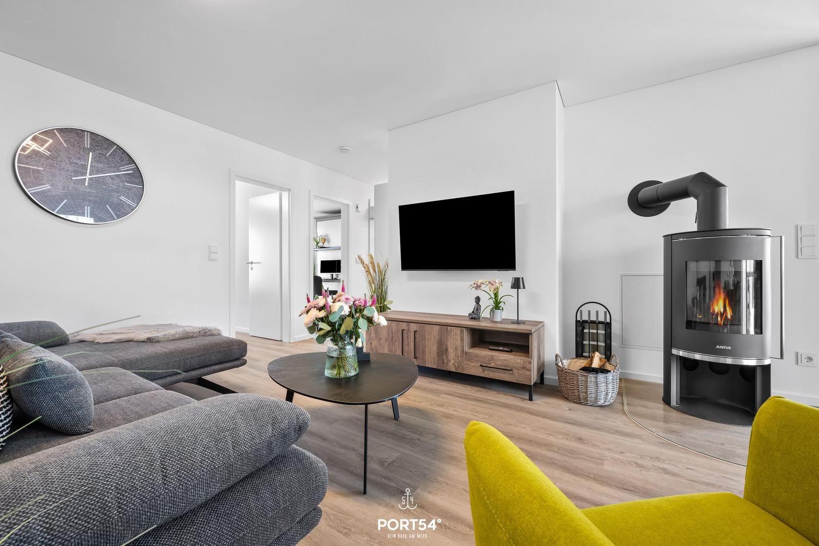 Wohnzimmer mit Sofa, Fernseher und Kaminofen. Holzfußboden und große Uhr an der Wand.