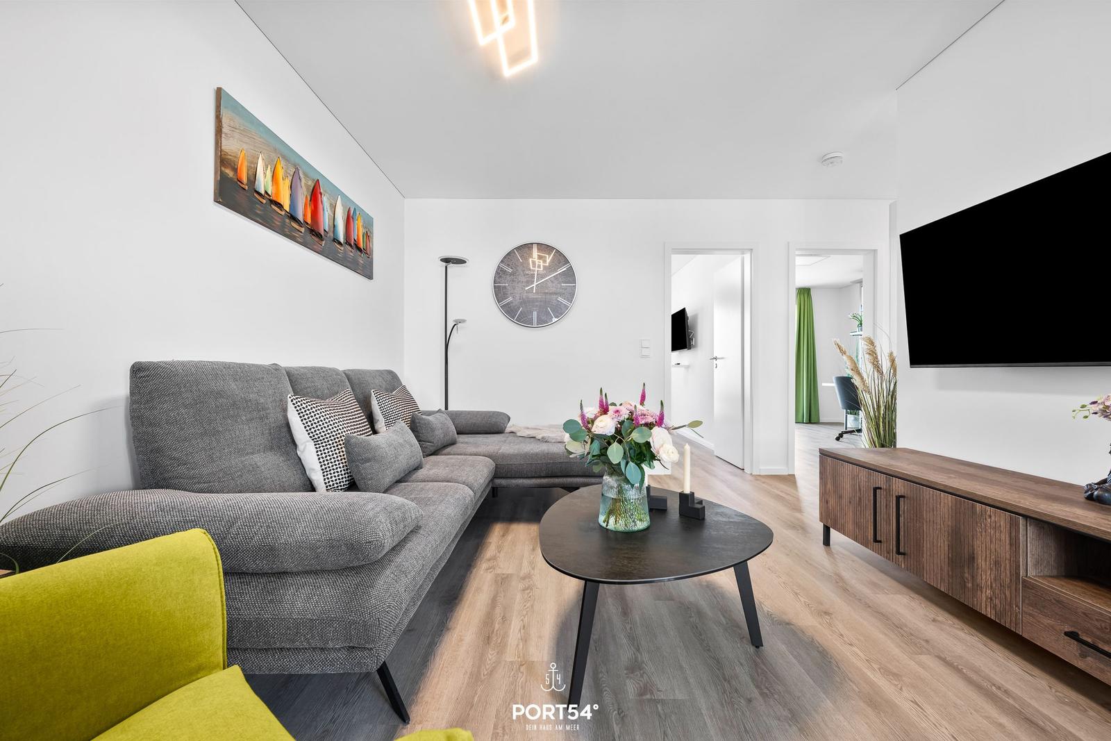 Wohnzimmer mit grauem Sofa, Holzboden und Wanduhr. Fernseher und Couchtisch sichtbar.