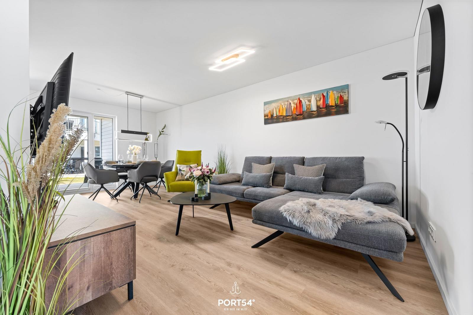 Wohn- und Essbereich mit grauem Sofa, grüner Sitzgelegenheit und Wandbild mit Segelbooten.