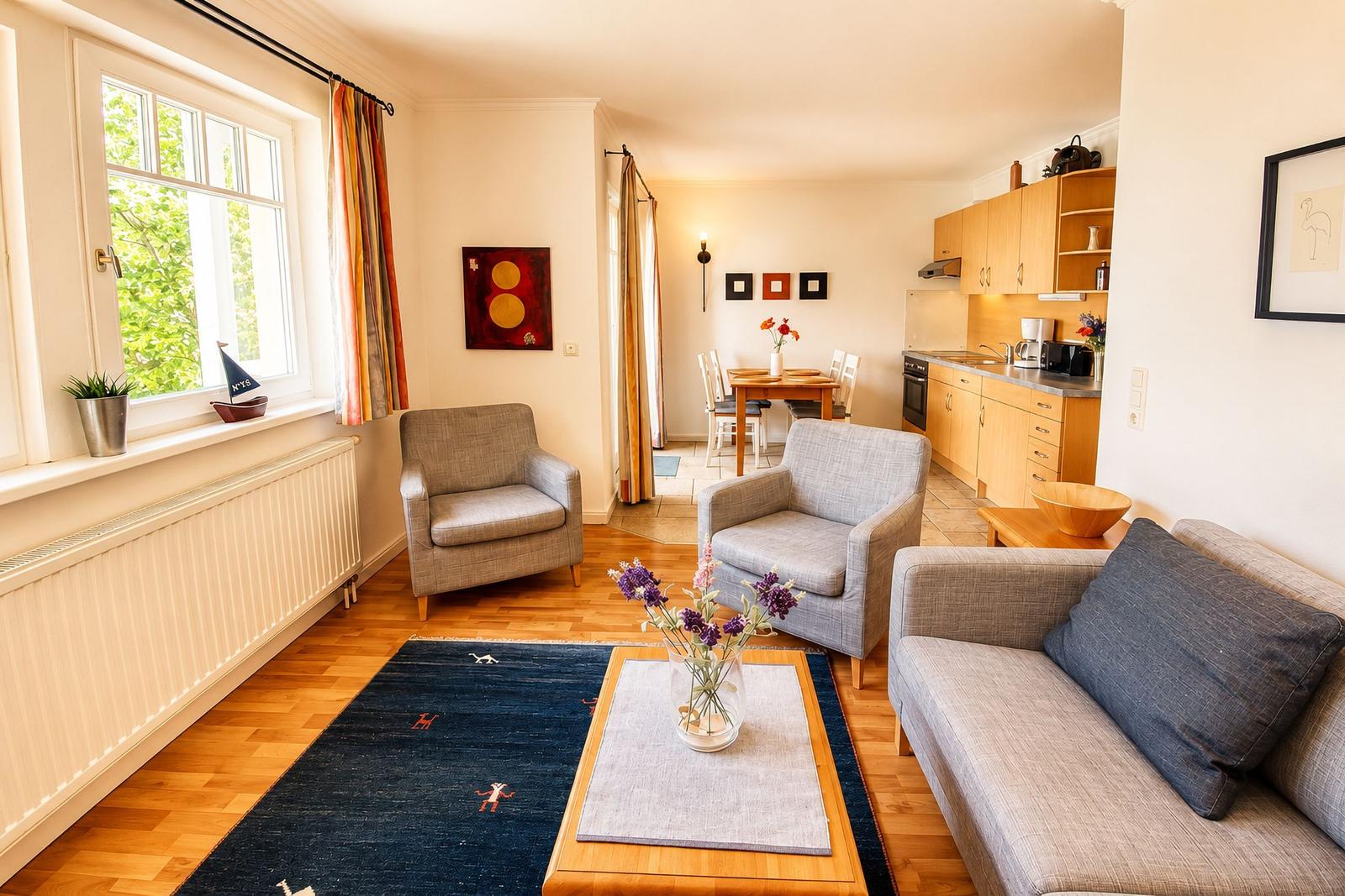 Wohnzimmer mit Sofa, zwei Sesseln, Tisch und Teppich. Küche und Essbereich im Hintergrund.