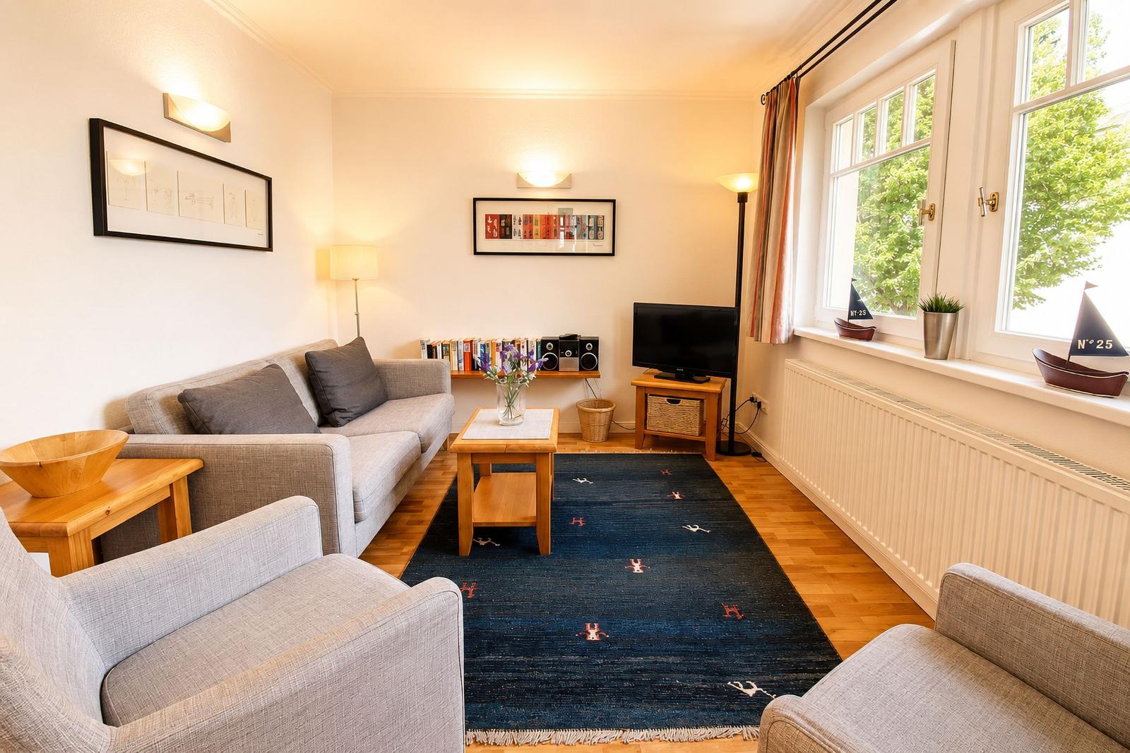 Wohnzimmer mit Sofa, Sesseln, Teppich und TV. Fenster mit Blick auf Grün.