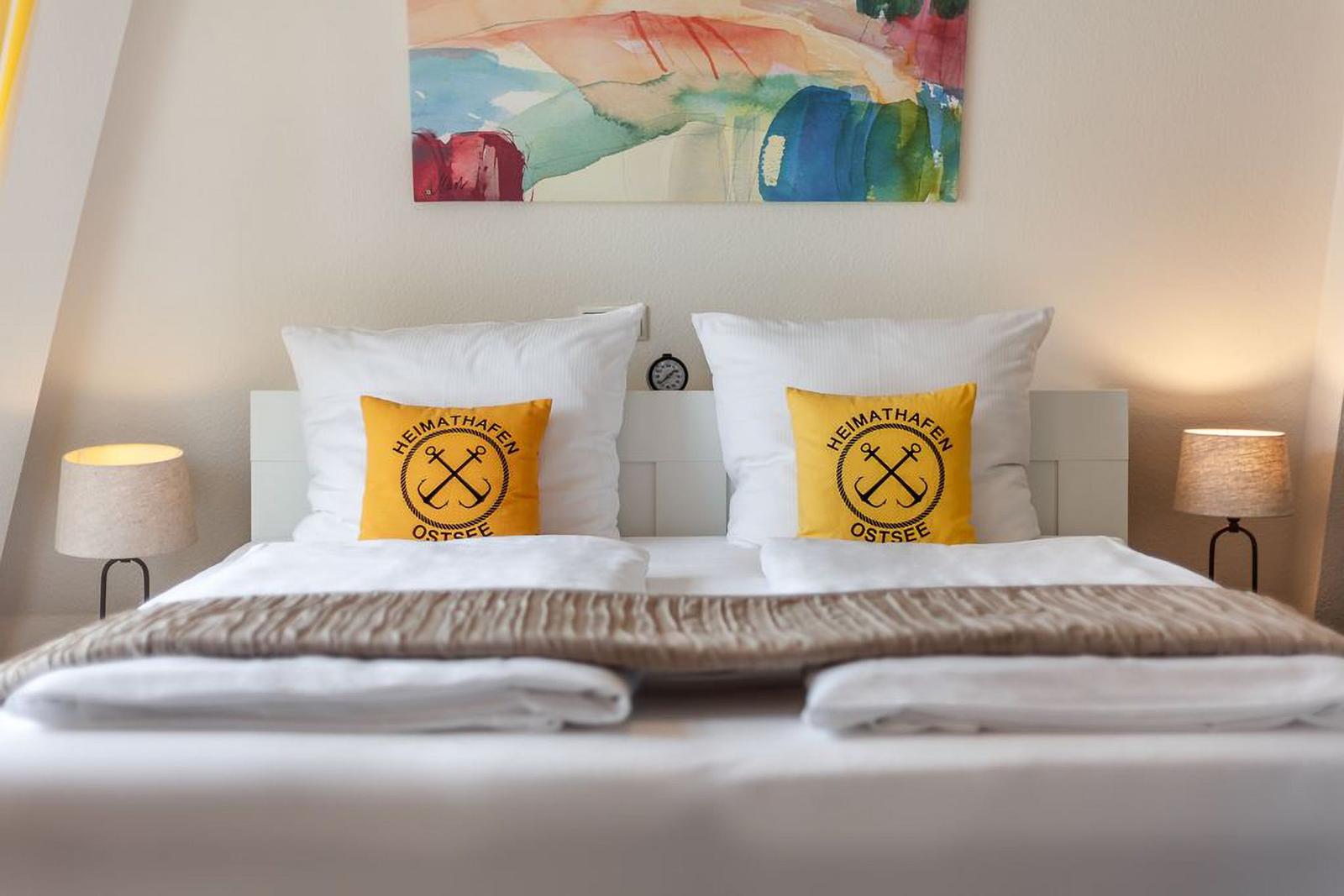 Ein Bett mit weißen Kissen und zwei gelben Kissen mit Logo. Über dem Bett hängt ein buntes Bild.