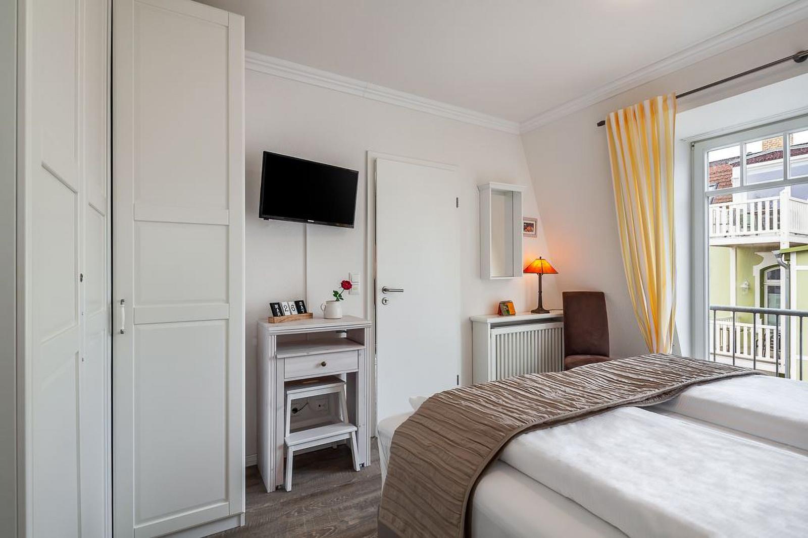 Schlafzimmer mit Bett, Schrank, Fernseher und Fenster mit Blick auf Balkon.