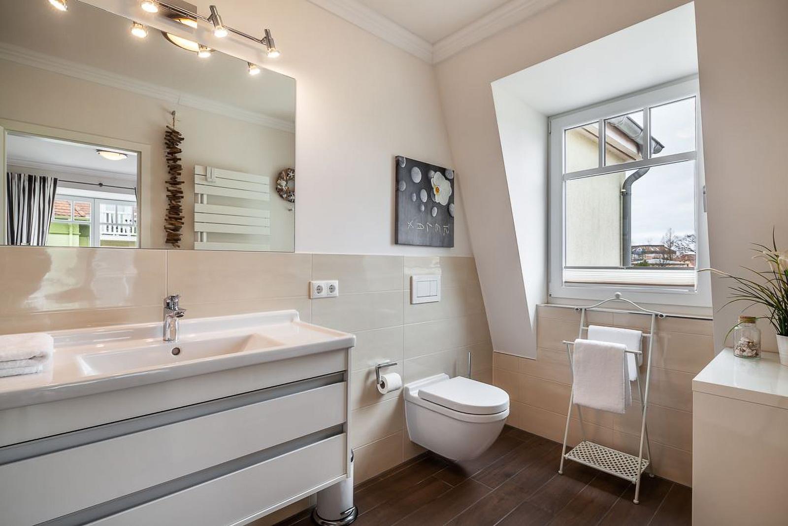 Badezimmer mit Waschbecken, Toilette und Fenster. Weiße Einrichtung und Holzboden.