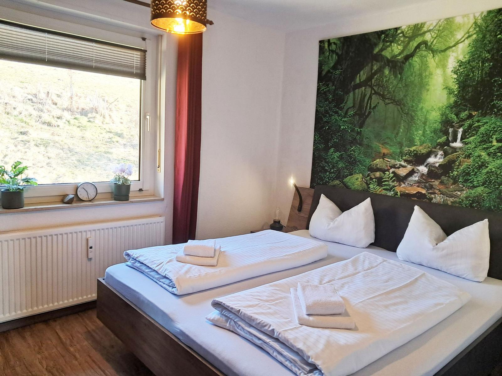 Schlafzimmer mit Bett, Fenster und Wandbild. Auf dem Bett liegen Handtücher.