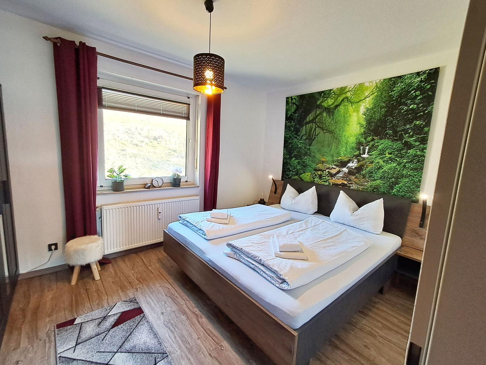 Schlafzimmer mit Doppelbett, Wandbild Wald, Fenster mit Blick, Holzboden und Deckenleuchte.