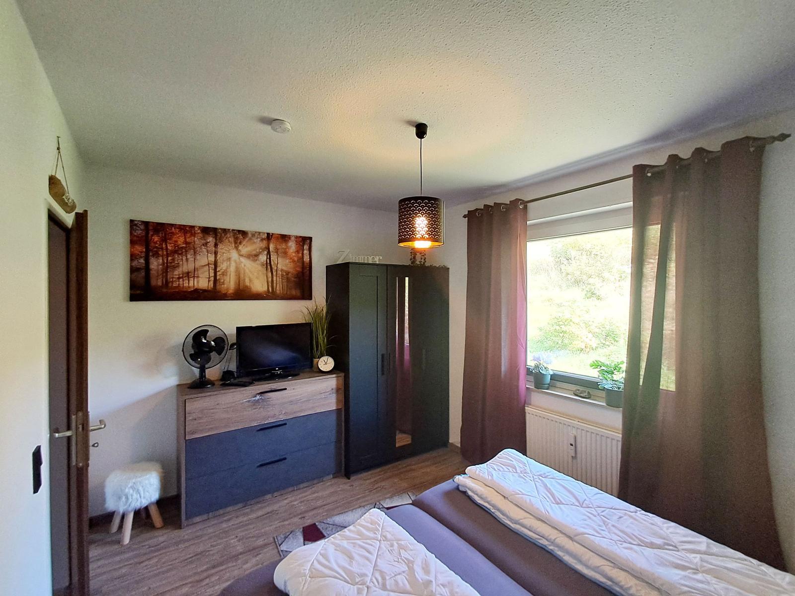Schlafzimmer mit Bett, Kleiderschrank, TV und Fenster mit Blick auf Grün.