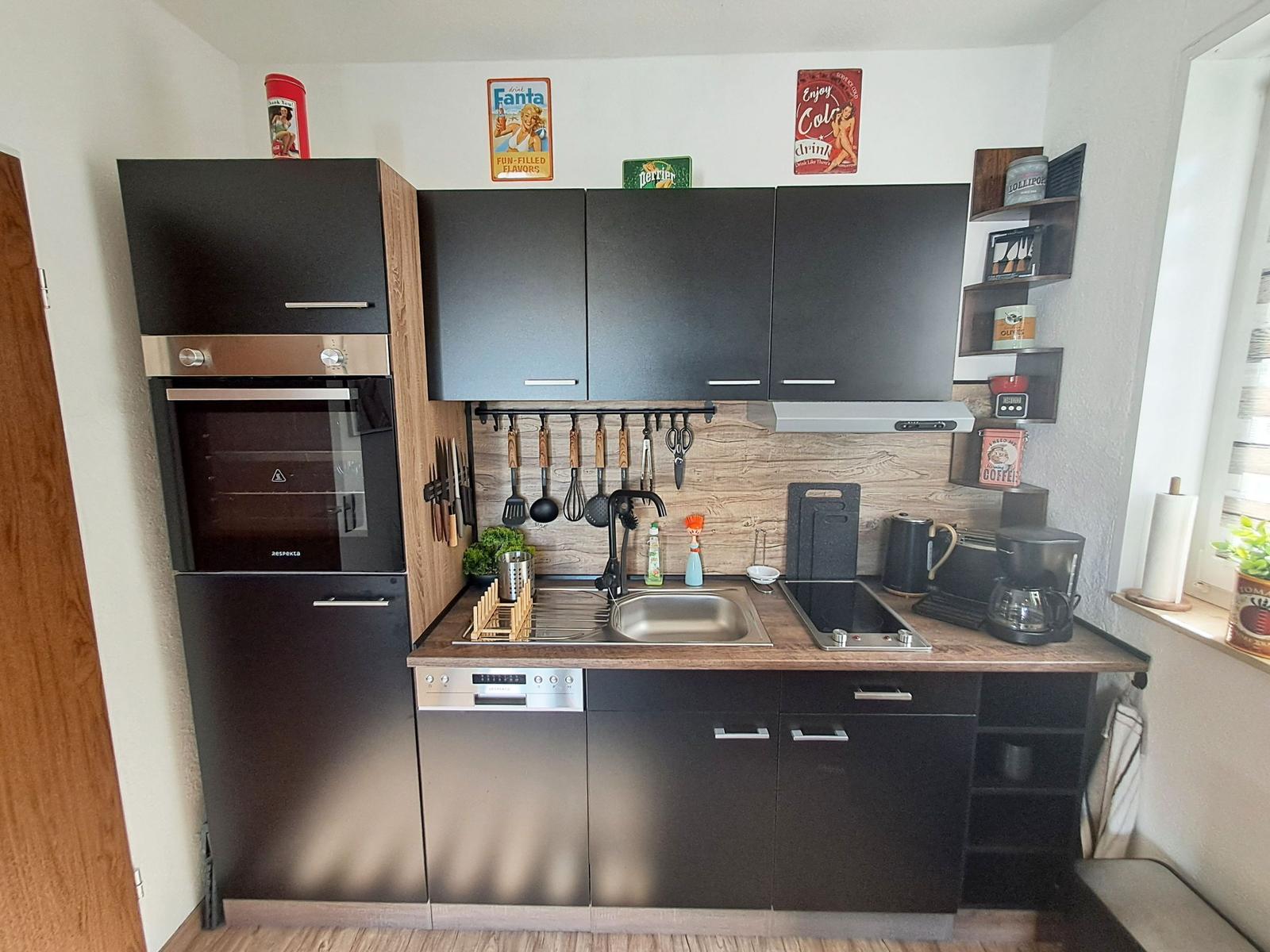 Küche mit schwarzen Schränken, Edelstahlspüle und Kochfeld. Dekorative Flaschen und Poster an der Wand.