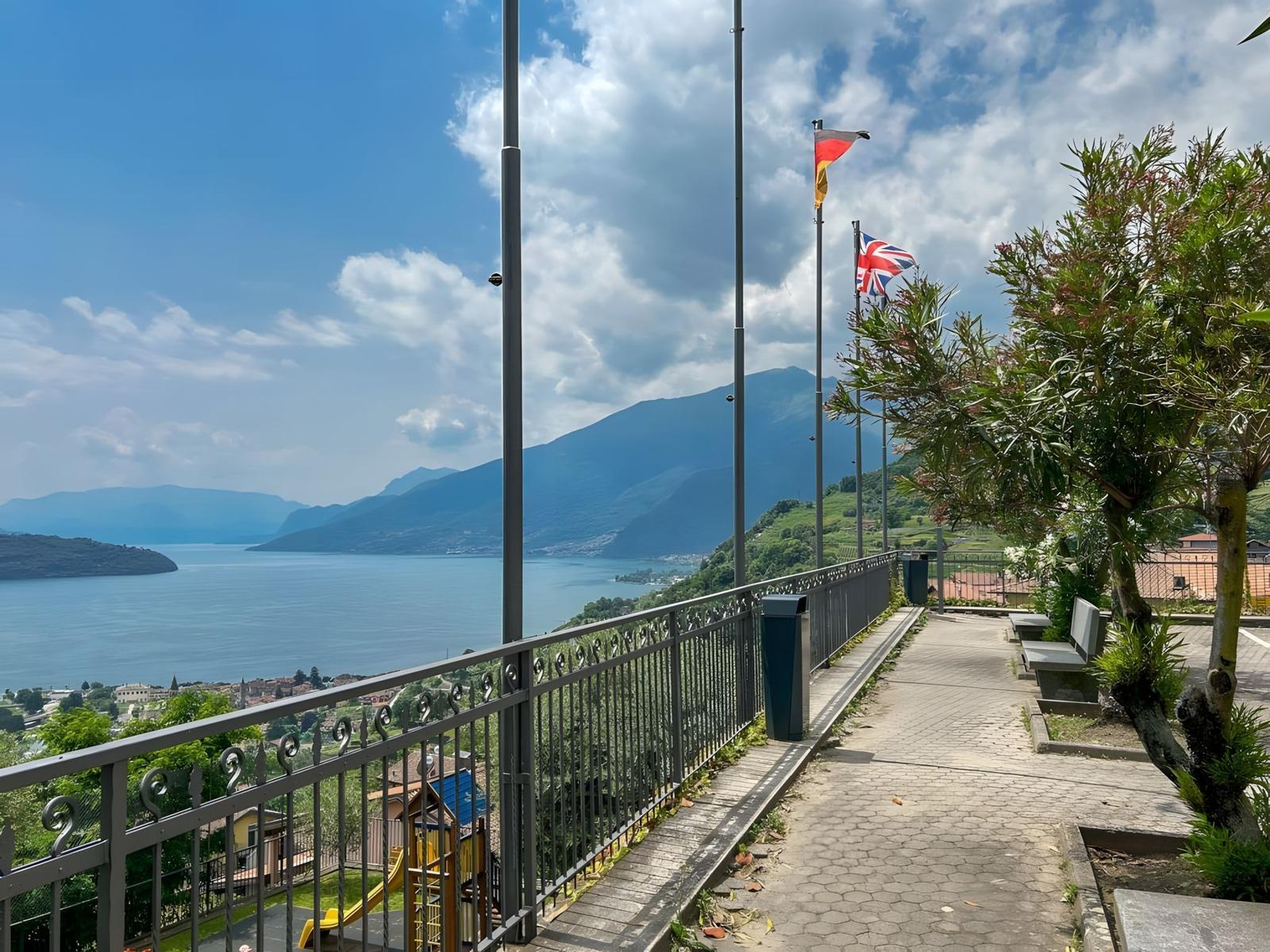 Weg mit Geländer und Flaggen überblickt den See und Berge.