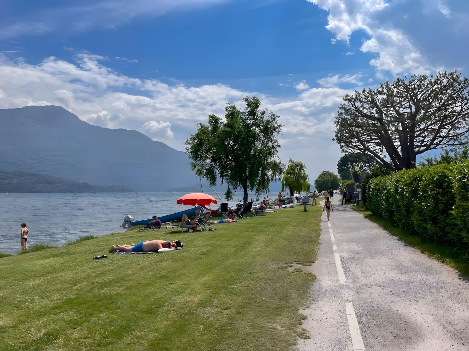 Grüner Strandabschnitt mit Sonnenbadenden, Booten und Bergen im Hintergrund.