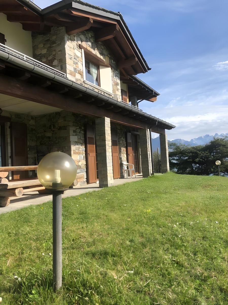 Steinhaus mit Holzverkleidung und Terrasse. Grasfläche und Berglandschaft im Hintergrund.