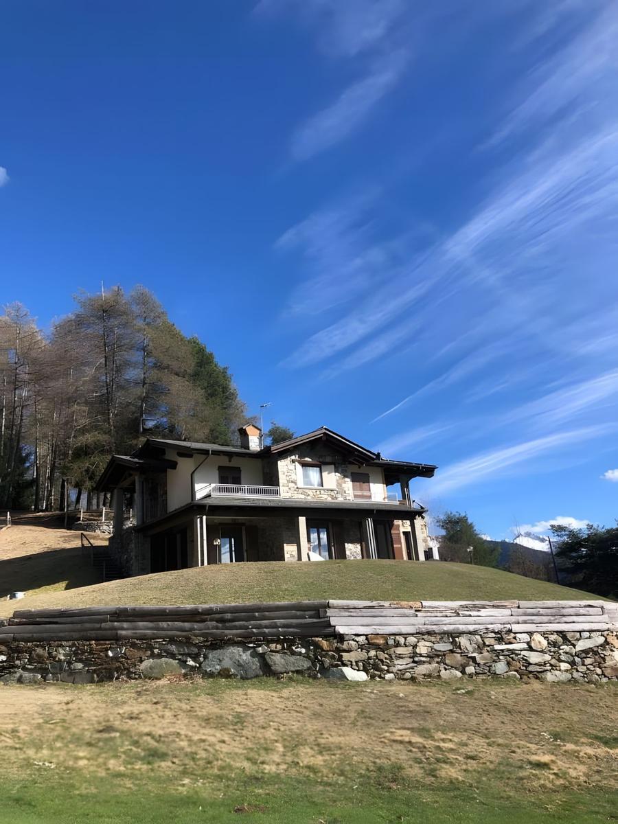Haus auf grüner Anhöhe mit Steinmauer und Wald im Hintergrund unter blauem Himmel.